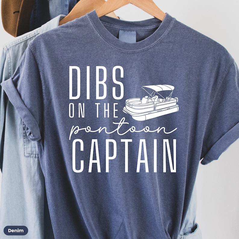 May include: A blue t-shirt with white text that reads "Dibs on the pontoon captain" and a white outline of a pontoon boat.