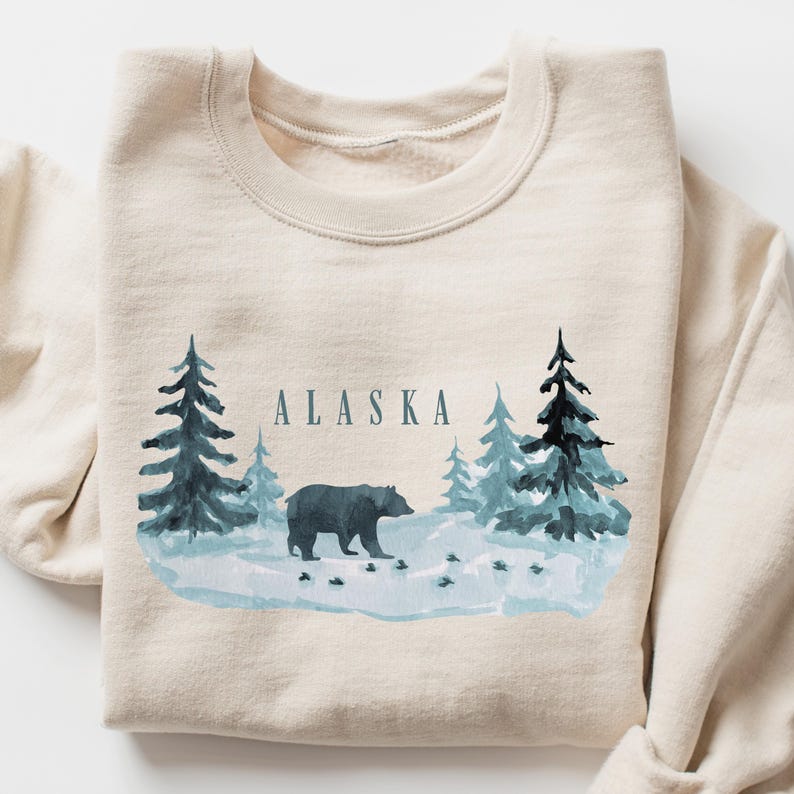May include: A beige sweatshirt with a watercolor design of a black bear walking through a snowy forest. The text "ALASKA" is printed in blue above the bear.