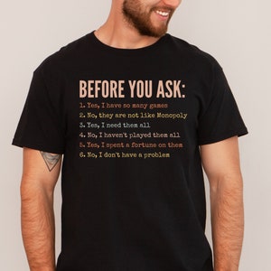 K&ouml;nnte beinhalten: Ein schwarzes T-Shirt mit wei&szlig;em Text, der "BEFORE YOU ASK:" lautet, gefolgt von einer Liste mit sechs Aussagen. Die Aussagen lauten: "1. Ja, ich habe so viele Spiele", "2. Nein, sie sind nicht wie Monopoly", "3. Ja, ich brauche sie alle", "4. Nein, ich habe nicht alle gespielt", "5. Ja, ich habe ein Verm&ouml;gen daf&uuml;r ausgegeben", und "6. Nein, ich habe kein Problem".