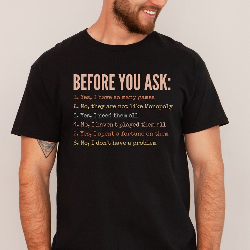 K&ouml;nnte beinhalten: Ein schwarzes T-Shirt mit wei&szlig;em Text, der "BEFORE YOU ASK:" lautet, gefolgt von einer Liste mit sechs Aussagen. Die Aussagen lauten: "1. Ja, ich habe so viele Spiele", "2. Nein, sie sind nicht wie Monopoly", "3. Ja, ich brauche sie alle", "4. Nein, ich habe nicht alle gespielt", "5. Ja, ich habe ein Verm&ouml;gen daf&uuml;r ausgegeben", und "6. Nein, ich habe kein Problem".