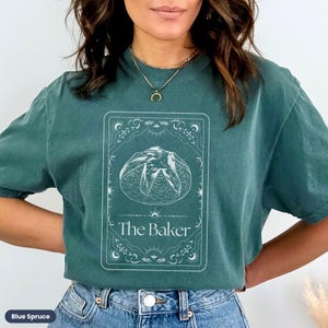 Pu&ograve; includere: Una maglietta verde acqua con una grafica bianca di una pagnotta di pane in una cornice decorativa. Il testo "The Baker" &egrave; stampato sotto il pane.