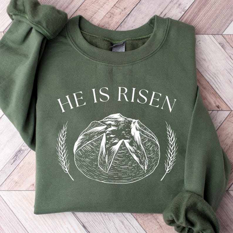May include: Olive green sweatshirt with the text "HE IS RISEN" in white. A white illustration of a loaf of bread with wheat stalks on either side is centered below the text. The sweatshirt has a crew neck and long sleeves.