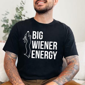 Pu&ograve; includere: Maglietta nera con una silhouette di bassotto bianca e il testo "BIG WIENER ENERGY" stampato sul davanti.