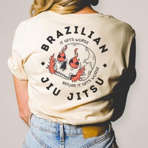 K&ouml;nnte beinhalten: Ein cremefarbenes T-Shirt mit einem schwarzen Kreis-Motiv auf der R&uuml;ckseite. Das Motiv zeigt einen Sch&auml;del mit Flammen als Augen und dem Text "Brazilian Jiu Jitsu" um den Sch&auml;del herum. Der Text im Kreis lautet "It gets worse before it gets worse."