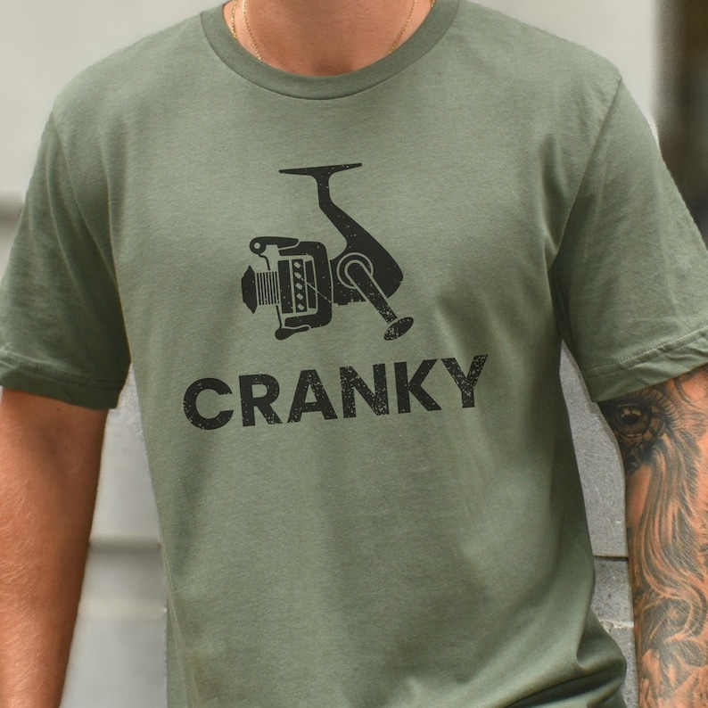 May include: A green t-shirt with a black graphic of a fishing reel and the word "CRANKY" printed on the front.