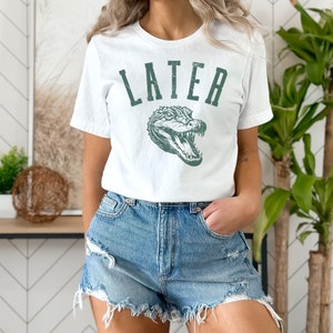 May include: White t-shirt with a green alligator graphic and the word "LATER" printed in a vintage style font.