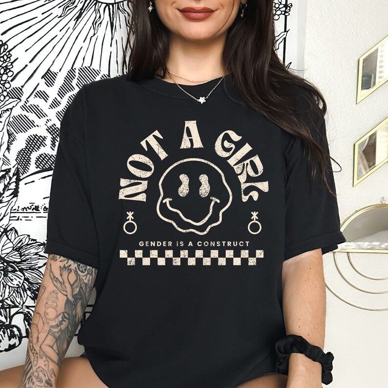 May include: Black t-shirt with a white graphic of a smiley face and the text "NOT A GIRL" in a circle. The text "GENDER IS A CONSTRUCT" is below the circle, and a checkered pattern is below the text.