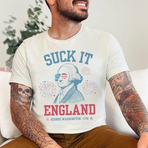 May include: White t-shirt with a blue and red graphic of George Washington wearing sunglasses and the text "SUCK IT ENGLAND GEORGE WASHINGTON, 1776".