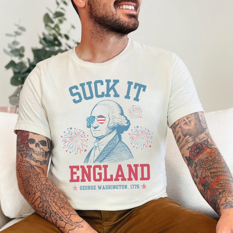 May include: White t-shirt with a blue and red graphic of George Washington wearing sunglasses and the text "SUCK IT ENGLAND GEORGE WASHINGTON, 1776".