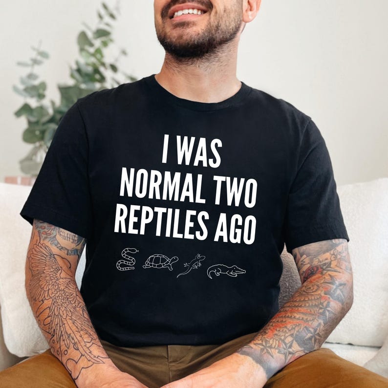 May include: A black t-shirt with white text that reads "I was normal two reptiles ago". The text is surrounded by small white illustrations of a snake, turtle, lizard, and alligator.