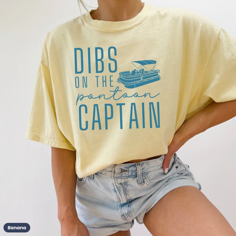 May include: A yellow t-shirt with the text "DIBS ON THE pontoon CAPTAIN" in blue lettering and a blue illustration of a pontoon boat.