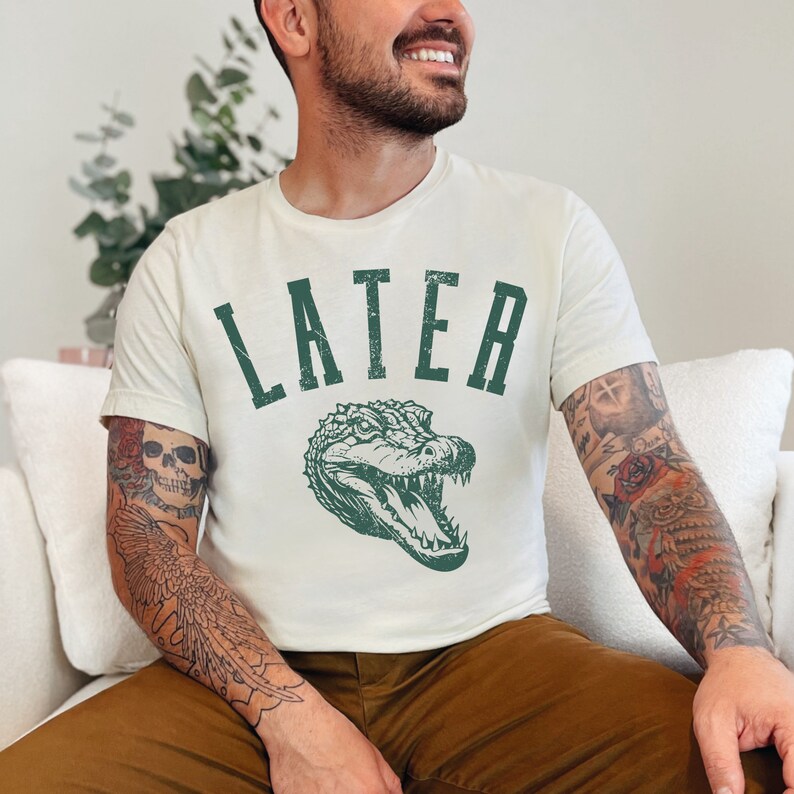 May include: A white t-shirt with a green alligator graphic and the word "LATER" printed in green. The person wearing the shirt has tattoos on their arms.