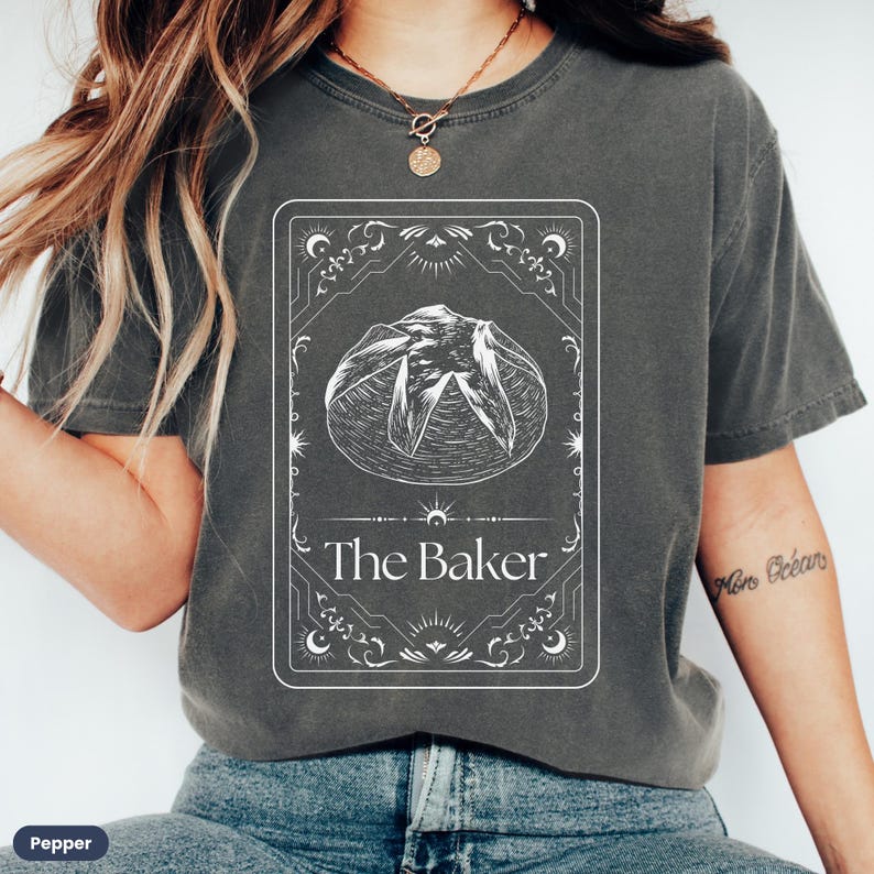 Pu&ograve; includere: Una maglietta grigia scura con una grafica bianca di una pagnotta di pane e il testo "The Baker" in una cornice decorativa.