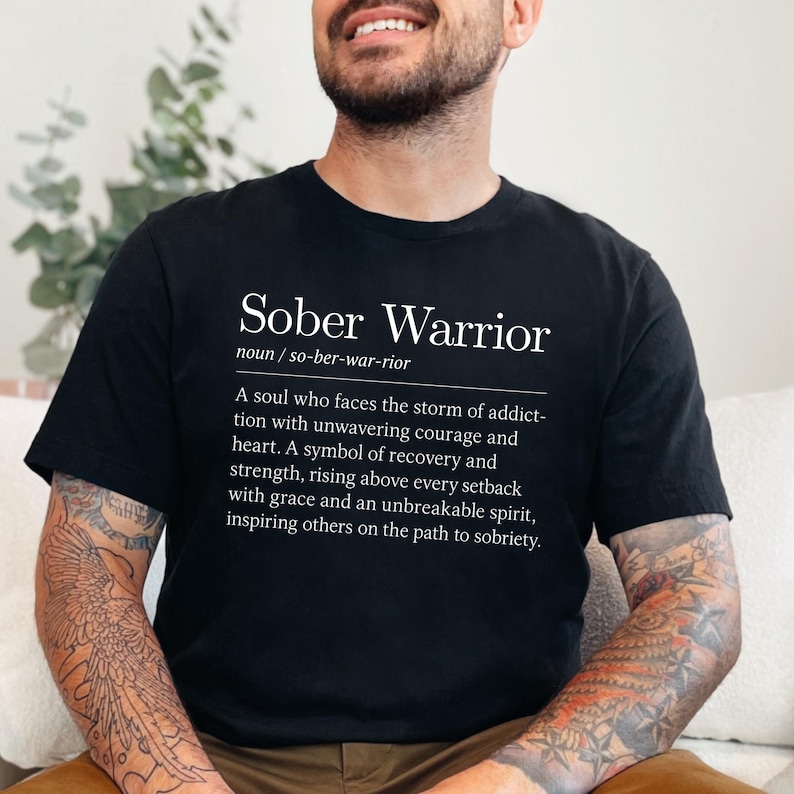 May include: Black t-shirt with white text that reads "Sober Warrior" and a definition of the term. The definition reads: "A soul who faces the storm of addiction with unwavering courage and heart. A symbol of recovery and strength, rising above every setback with grace and an unbreakable spirit, inspiring others on the path to sobriety."