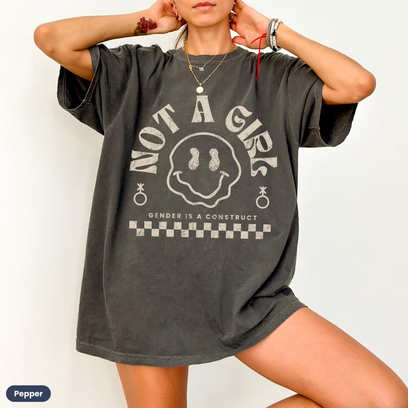 May include: A dark gray oversized t-shirt with a graphic print of a smiley face and the text "Not a Girl" in a retro font. The text "Gender is a construct" is printed below the graphic.