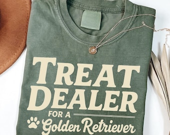 Puede incluir: Una camiseta verde con el texto "Treat Dealer for a Golden Retriever" en color crema. La camiseta tiene un aspecto desgastado y un gráfico de huella de pata.