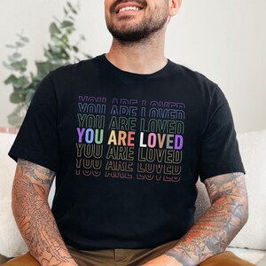 K&ouml;nnte beinhalten: Schwarzes T-Shirt mit dem Text "YOU ARE LOVED", der in Regenbogenfarben wiederholt wird.