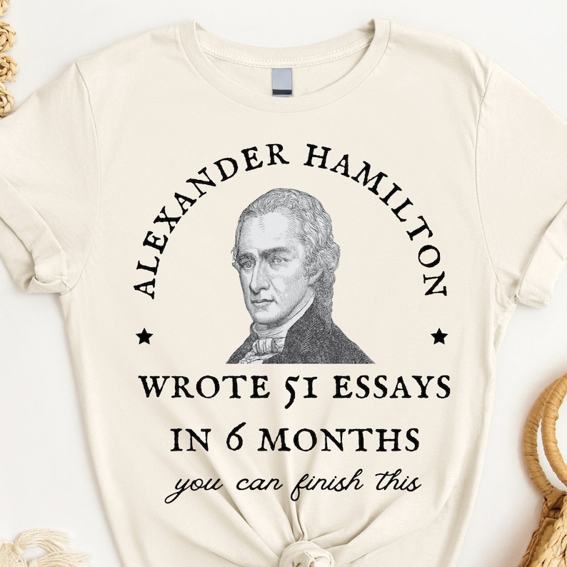 May include: A beige t-shirt with a black graphic of Alexander Hamilton. The graphic includes a portrait of Hamilton and the text "Wrote 51 essays in 6 months you can finish this".
