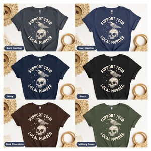May include: Six different color t-shirts with the text "Support Your Local Murder" and a graphic of a skull with a raven perched on top. The colors are dark heather, navy, black, dark chocolate, and military green.