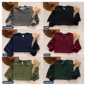 May include: A collection of crewneck sweatshirts in various colors, including dark heather, black, navy, maroon, military green, and forest green. Each sweatshirt is displayed with a hat and a bundle of wheat.