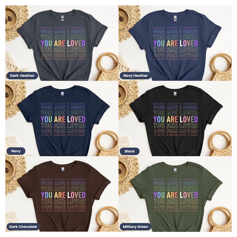 K&ouml;nnte beinhalten: Sechs T-Shirts in verschiedenen Farben mit dem Text "You Are Loved", der in einem Regenbogen-Farbverlauf wiederholt wird. Die Farben der T-Shirts sind dunkelgrau meliert, marineblau meliert, marineblau, schwarz, dunkelbraun und milit&auml;rgr&uuml;n.
