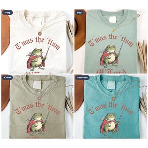May include: Four different colored t-shirts with a graphic print of a frog wearing a red cape and holding a sword. The text on the shirts reads "T'was the tism". The shirts are available in ivory, khaki, bay, and seafoam.