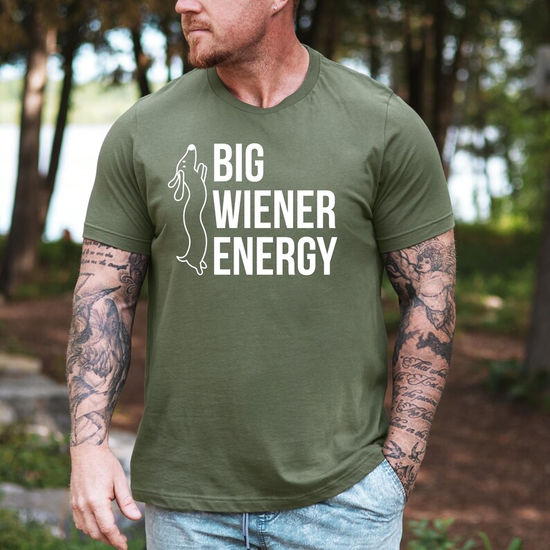 Pu&ograve; includere: Una maglietta verde con una silhouette di bassotto bianca e il testo "BIG WIENER ENERGY".