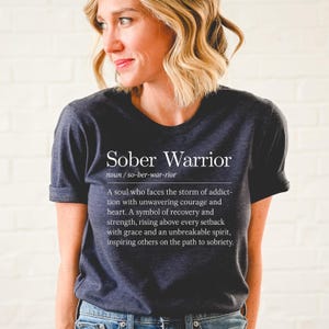 May include: A dark gray t-shirt with the text "Sober Warrior" and a definition of the term. The definition reads: "A soul who faces the storm of addiction with unwavering courage and heart. A symbol of recovery and strength, rising above every setback with grace and an unbreakable spirit, inspiring others on the path to sobriety."