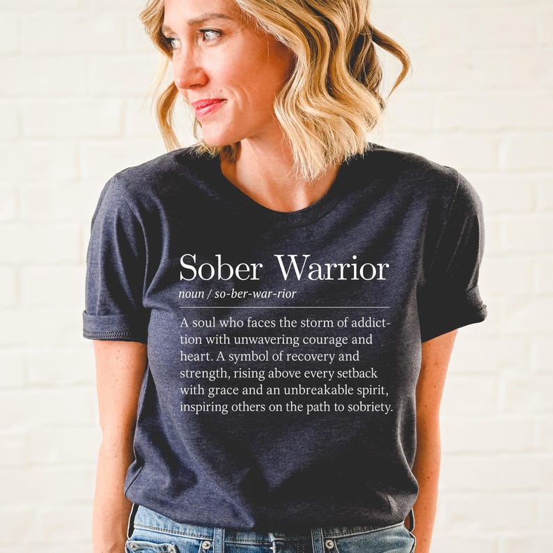 May include: A dark gray t-shirt with the text "Sober Warrior" and a definition of the term. The definition reads: "A soul who faces the storm of addiction with unwavering courage and heart. A symbol of recovery and strength, rising above every setback with grace and an unbreakable spirit, inspiring others on the path to sobriety."