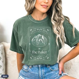 Pu&ograve; includere: Una maglietta verde con una grafica bianca di una pagnotta di pane in una cornice decorativa. Il testo "The Baker" &egrave; sotto il pane.