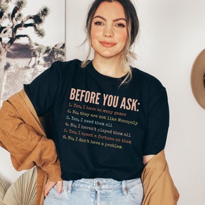 K&ouml;nnte beinhalten: Ein schwarzes T-Shirt mit wei&szlig;em Text, der "BEFORE YOU ASK:" lautet, gefolgt von einer Liste mit sechs Aussagen. Die Aussagen lauten: "1. Ja, ich habe so viele Spiele", "2. Nein, sie sind nicht wie Monopoly", "3. Ja, ich brauche sie alle", "4. Nein, ich habe nicht alle gespielt", "5. Ja, ich habe ein Verm&ouml;gen daf&uuml;r ausgegeben", und "6. Nein, ich habe kein Problem".