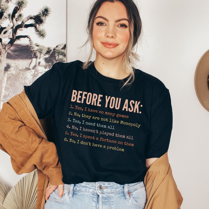 K&ouml;nnte beinhalten: Ein schwarzes T-Shirt mit wei&szlig;em Text, der "BEFORE YOU ASK:" lautet, gefolgt von einer Liste mit sechs Aussagen. Die Aussagen lauten: "1. Ja, ich habe so viele Spiele", "2. Nein, sie sind nicht wie Monopoly", "3. Ja, ich brauche sie alle", "4. Nein, ich habe nicht alle gespielt", "5. Ja, ich habe ein Verm&ouml;gen daf&uuml;r ausgegeben", und "6. Nein, ich habe kein Problem".