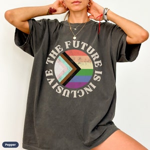 May include: Dark grey t-shirt with a circular graphic that reads "THE FUTURE IS INCLUSIVE." The graphic features a rainbow and other pride flag designs. The shirt is a relaxed fit.