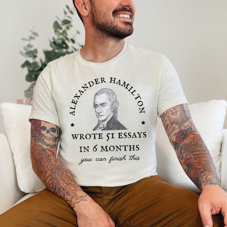 May include: A white t-shirt with a black graphic of Alexander Hamilton, a quote that reads "Wrote 51 essays in 6 months you can finish this", and a black star on each side of the quote.