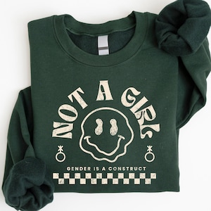 May include: A dark green sweatshirt with a white graphic print that says "Not a Girl" with a smiley face and the text "Gender is a construct" below. The graphic is surrounded by a checkered pattern.