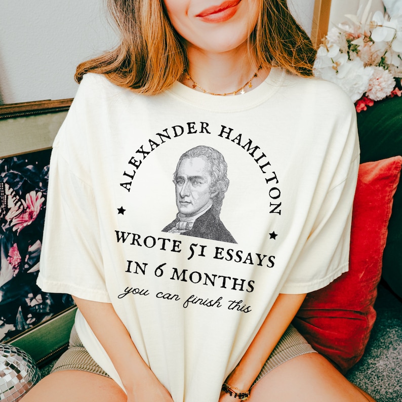 May include: A white t-shirt with a black graphic of Alexander Hamilton. The graphic includes a portrait of Hamilton and the text "Alexander Hamilton wrote 51 essays in 6 months you can finish this".