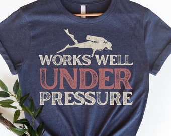 May include: A navy blue t-shirt with a white silhouette of a scuba diver and the text "WORKS WELL UNDER PRESSURE" in white and red.