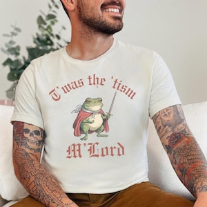 May include: A white t-shirt with a graphic of a frog wearing a red cape and holding a sword. The text "T'was the 'tism M'Lord" is printed on the shirt.