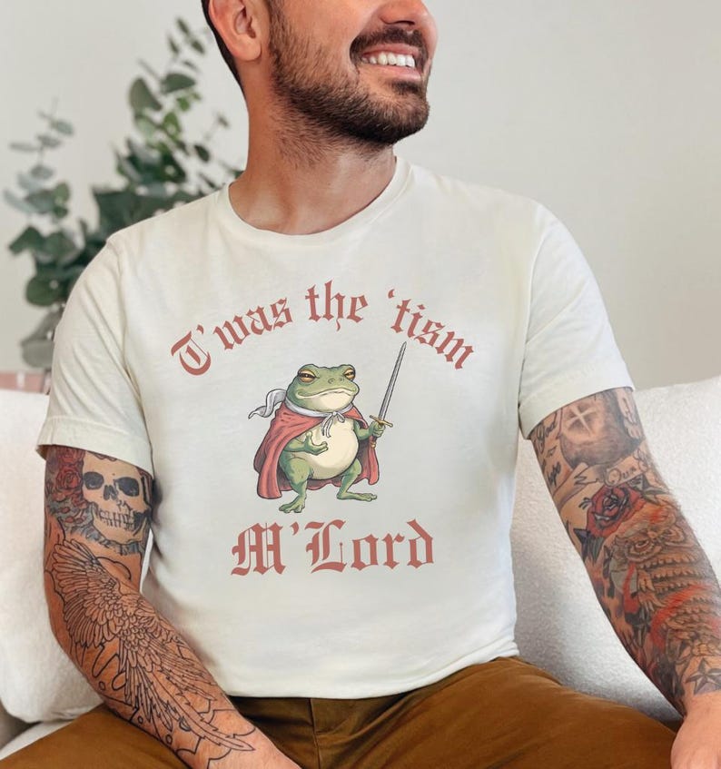 May include: A white t-shirt with a graphic of a frog wearing a red cape and holding a sword. The text "T'was the 'tism M'Lord" is printed on the shirt.
