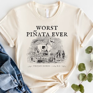 以下が含まれることがあります： 「WORST PI&Ntilde;ATA EVER」の文字と、トロイの木馬の白黒イラストがプリントされたクリーム色のTシャツ。「TROJAN HORSE - 1184 B.C.」の文字が下にあります。