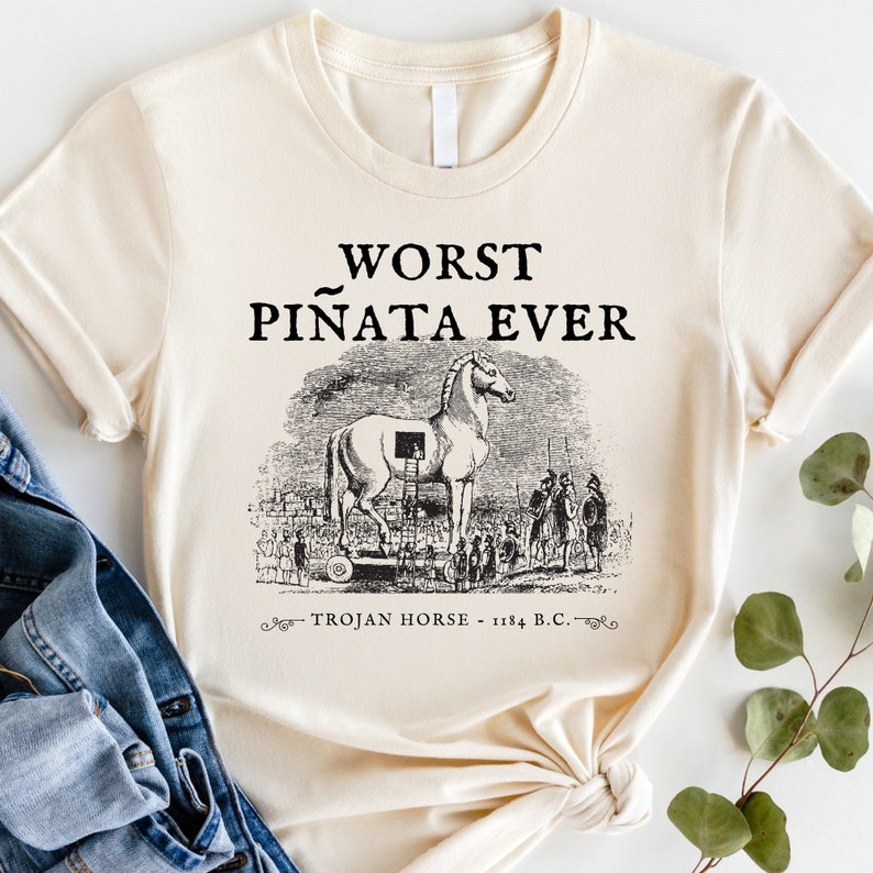 以下が含まれることがあります： 「WORST PI&Ntilde;ATA EVER」の文字と、トロイの木馬の白黒イラストがプリントされたクリーム色のTシャツ。「TROJAN HORSE - 1184 B.C.」の文字が下にあります。
