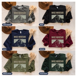 May include: A set of six sweatshirts in various colors, each featuring a vintage-style graphic of a mountain range with the text "ROCKY MOUNTAINS COLORADO". The colors include dark heather, navy, military green, black, maroon, and forest green.
