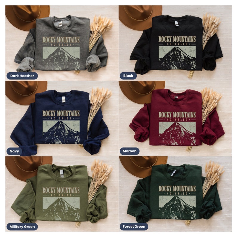 May include: A set of six sweatshirts in various colors, each featuring a vintage-style graphic of a mountain range with the text "ROCKY MOUNTAINS COLORADO". The colors include dark heather, navy, military green, black, maroon, and forest green.