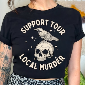 May include: Black cropped t-shirt with a white graphic design. The graphic features a skull with a crow perched on top. The text "Support Your Local Murder" is written around the skull and crow.