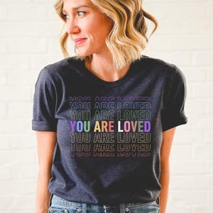 K&ouml;nnte beinhalten: Ein dunkel-melierter grauer T-Shirt mit dem Text "YOU ARE LOVED", der in Regenbogenfarben wiederholt wird.