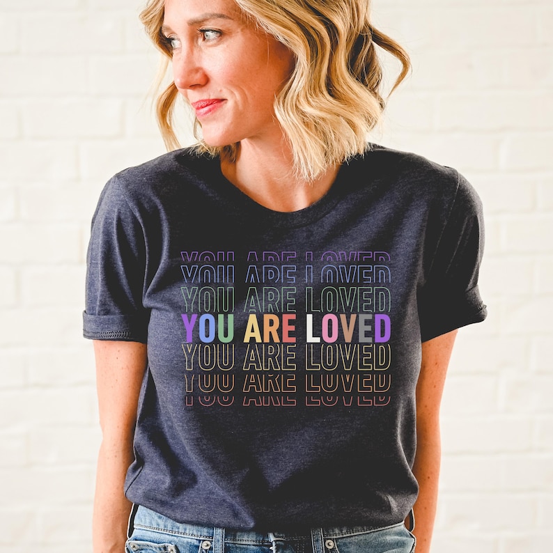 K&ouml;nnte beinhalten: Ein dunkel-melierter grauer T-Shirt mit dem Text "YOU ARE LOVED", der in Regenbogenfarben wiederholt wird.