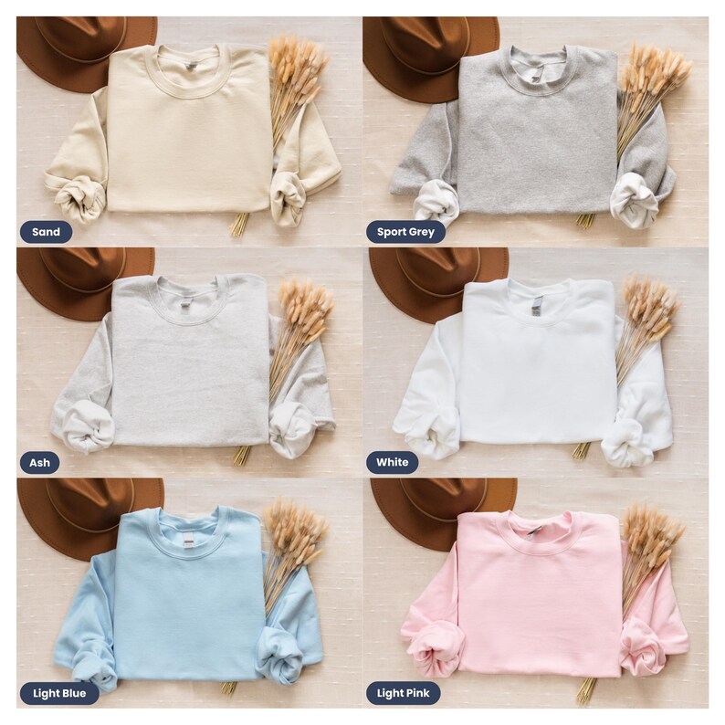 May include: Six different color sweatshirts laid flat on a light brown surface. The sweatshirts are all the same style, with long sleeves and a crew neck. The colors are sand, sport grey, ash, white, light blue, and light pink.