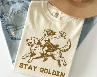 Puede incluir: Una camiseta de color crema con una ilustración marrón de una mujer montando un golden retriever. El texto "STAY GOLDEN" está impreso en marrón en la camiseta.