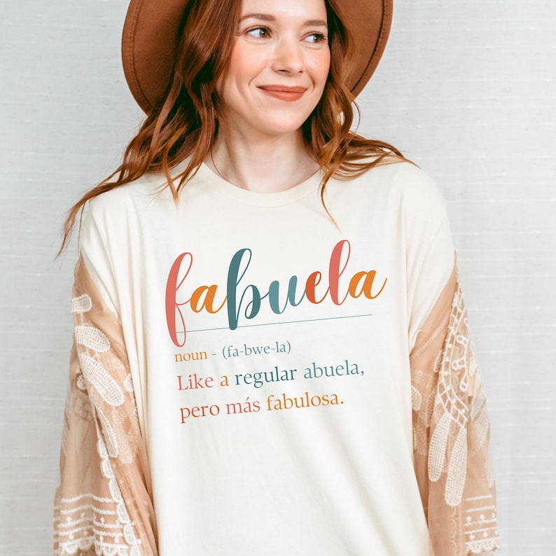 Puede incluir: Una camiseta blanca con la palabra "fabuela" escrita en letras de colores. El texto debajo define "fabuela" como un sustantivo, que significa "como una abuela normal, pero m&aacute;s fabulosa."