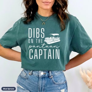 May include: A teal blue t-shirt with white text that reads "Dibs on the pontoon captain" and a white illustration of a pontoon boat.